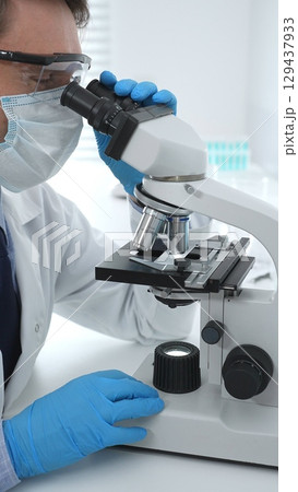 Man scientist wearing protective googles and blue gloves, is using microscope analyzing samples in modern laboratory doing scientific research. Medicine and science concept 129437933
