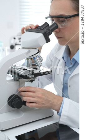 Female scientist wearing protective gear analyzing test samples under microscope in contemporary research environment. Medicine, healthcare and science concept Female scientist wearing protective gear analyzing test samples under microscope in contemporary research environment. Medicine, healthcare and science concept 129437937