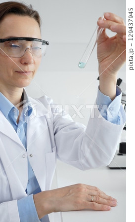 Female scientist wearing lab coat and protective glasses holding a test tube with blue liquid, conducting research in a modern laboratory setting. Medicine, health care and science concept Female scientist wearing lab coat and protective glasses holding a test tube with blue liquid, conducting research in a modern laboratory setting. Medicine, health care and science concept 129437945