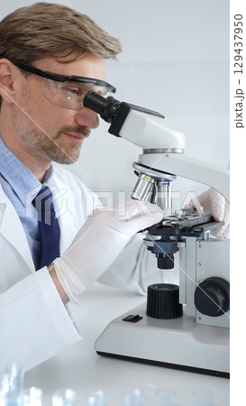 Researcher examining sample through microscope, wearing safety eyewear in sterile laboratory setting, surrounded by scientific glassware. Medicine, healthcare and science concept Researcher examining sample through microscope, wearing safety eyewear in sterile laboratory setting, surrounded by scientific glassware. Medicine, healthcare and science concept 129437950