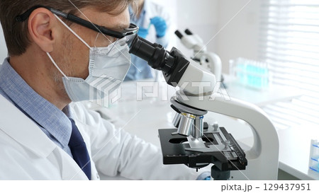 Medical researcher analyzing sample under microscope, wearing protective gear in sterile laboratory setting, performing scientific research. Medicine, healthcare and science concept 129437951