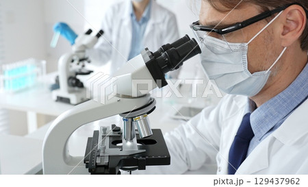 Masked scientist analyzing sample under microscope, colleague researching nearby in sterile medical laboratory setting. Medicine, healthcare and science concept Masked scientist analyzing sample under microscope, colleague researching nearby in sterile medical laboratory setting. Medicine, healthcare and science concept 129437962