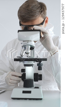 A male medical virologist in a medical gown and gloves examines viral samples under a microscope while conducting research in a sterile laboratory setting A male medical virologist in a medical gown and gloves examines viral samples under a microscope while conducting research in a sterile laboratory setting 129437977