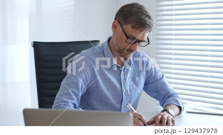 Male adult accountant wearing blue sleek shirt and black glasses is concentrating while working at the desk in modern office, demonstrating professionalism. Business people, taxes and audit concept Male adult accountant wearing blue sleek shirt and black glasses is concentrating while working at the desk in modern office, demonstrating professionalism. Business people, taxes and audit concept 129437978