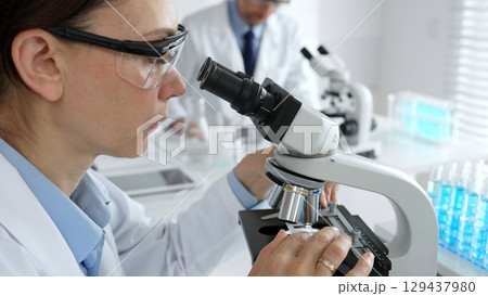 Female scientist examining specimen under microscope, conducting research in sterile laboratory with colleague working near test tube rack. Medicine, healthcare and science concept Female scientist examining specimen under microscope, conducting research in sterile laboratory with colleague working near test tube rack. Medicine, healthcare and science concept 129437980