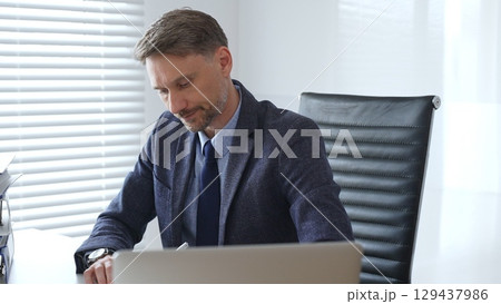 Adult businessman wearing blue suit and tie. is sitting at a modern desk, working intently on a laptop surrounded by folders, showcasing professionalism and determination. Audit and taxes in business Adult businessman wearing blue suit and tie. is sitting at a modern desk, working intently on a laptop surrounded by folders, showcasing professionalism and determination. Audit and taxes in business 129437986
