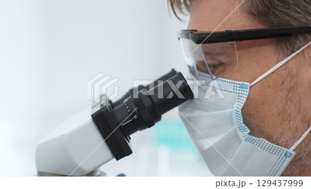 Close up of a scientist researching with a microscope, wearing protective glasses and a mask in a laboratory setting. Medicine, healthcare and science concept 129437999