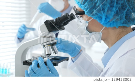 Close up of woman scientist wearing blue protective gear while examining specimen under microscope, laboratory assistant handles glassware in medical investigation at the background. Medicine concept 129438002