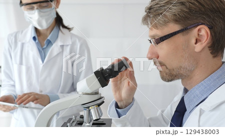 Scientific professional examining specimen through microscope, laboratory assistant nearby wearing protective mask, both dressed in white lab coats during research process. Medicine and science 129438003
