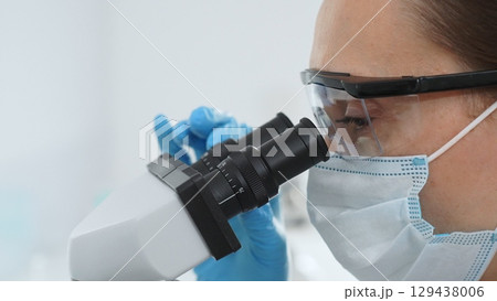 Close up of female scientist wearing safety glasses looking through microscope, conducting important research in modern laboratory setting. Medicine, healthcare and science concept Close up of female scientist wearing safety glasses looking through microscope, conducting important research in modern laboratory setting. Medicine, healthcare and science concept 129438006
