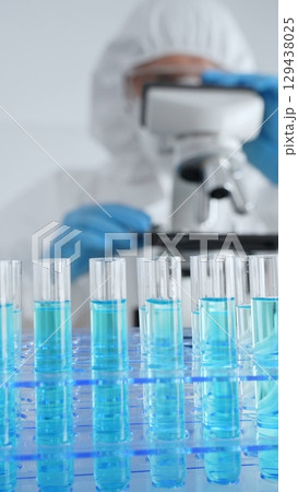 Close up of test tubes with blue liquid opposite a scientific researcher wearing protective gear tuning microscope lenses in laboratory. Medicine, healthcare and science concept Close up of test tubes with blue liquid opposite a scientific researcher wearing protective gear tuning microscope lenses in laboratory. Medicine, healthcare and science concept 129438025
