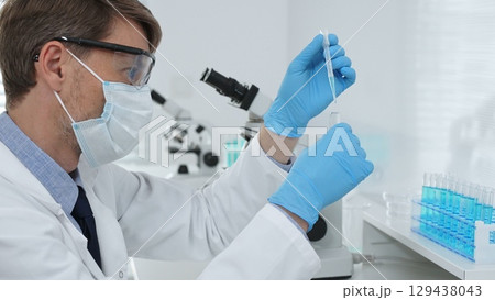 Masked male scientist in lab coat, protective glasses and blue gloves is adding reagent to test tube with microscopes and test tube rack in modern laboratory. Medicine, healthcare and science concept 129438043
