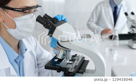Female scientist wearing mask and gloves using microscope doing scientific research in modern laboratory with colleague working in background. Medicine, healthcare and science concept Female scientist wearing mask and gloves using microscope doing scientific research in modern laboratory with colleague working in background. Medicine, healthcare and science concept 129438046