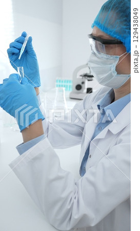 Medical science researcher wearing blue protective gear of cap, gloves and googles, is examining liquid in test tube in laboratory workspace. Medicine and health care concept Medical science researcher wearing blue protective gear of cap, gloves and googles, is examining liquid in test tube in laboratory workspace. Medicine and health care concept 129438049