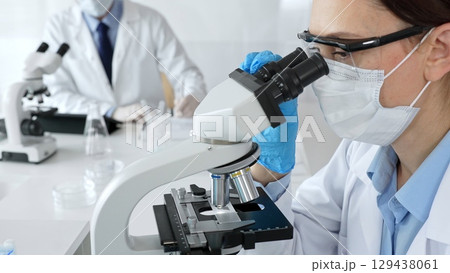 A female scientist in a blue mask and gloves is using a microscope and conducting scientific research in a sterile laboratory while a colleague is working in the background. Medicine, healthcare and A female scientist in a blue mask and gloves is using a microscope and conducting scientific research in a sterile laboratory while a colleague is working in the background. Medicine, healthcare and 129438061