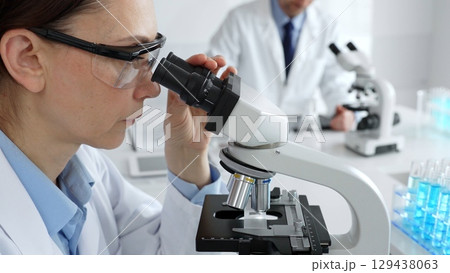 Female scientist examining specimen under microscope, conducting research in sterile laboratory with colleague working near test tube rack. Medicine, healthcare and science concept 129438063