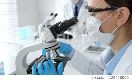 A female scientist in a blue mask and gloves is using a microscope and conducting scientific research in a sterile laboratory while a colleague is working in the background. Medicine, healthcare and A female scientist in a blue mask and gloves is using a microscope and conducting scientific research in a sterile laboratory while a colleague is working in the background. Medicine, healthcare and 129438066