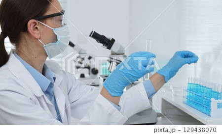 Female scientist wearing a face mask and blue gloves, carefully dropping liquid from a pipette into a test tube in a modern lab. Medicine, healthcare and science concept Female scientist wearing a face mask and blue gloves, carefully dropping liquid from a pipette into a test tube in a modern lab. Medicine, healthcare and science concept 129438070