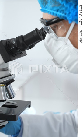 Medical scientist senior man wearing protective mask, glasses and bouffant cap using microscope doing scientific research in a laboratory. Medicine and science concept 129438112