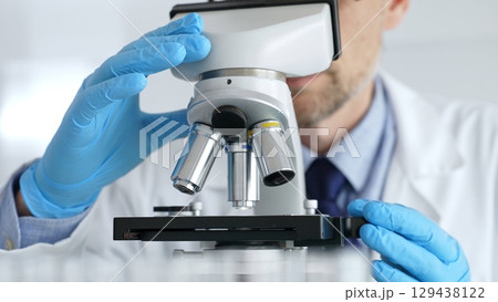 Male scientist in white coat and blue gloves adjusting microscope, conducting pharmaceutical research in modern laboratory. Medicine, healthcare and science concept Male scientist in white coat and blue gloves adjusting microscope, conducting pharmaceutical research in modern laboratory. Medicine, healthcare and science concept 129438122