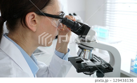Female scientist examining microscope slide, analyzing research data in sterile laboratory surrounded by scientific glassware and equipment. Medicine, healthcare and science concept 129438136