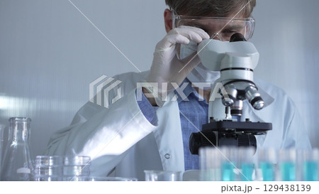 Male scientist, wearing a lab coat, mask, white gloves and safety glasses, is using a binocular microscope surrounded by test tubes in laboratory 129438139