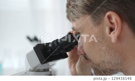 Close up of a man scientist carefully observing through a microscope in a brightly lit laboratory, conducting research for developing new medicines and treatments. Medicine and science concept 129438148