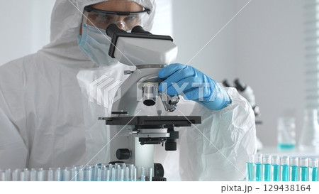 Medical virologist in protective hazmat gear examining viral samples under a microscope while conducting research in a sterile laboratory environment Medical virologist in protective hazmat gear examining viral samples under a microscope while conducting research in a sterile laboratory environment 129438164