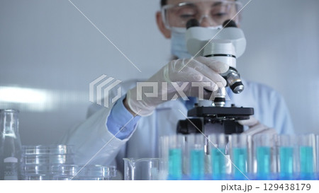 A scientist woman, wearing a lab coat, mask, white gloves and safety glasses, is adjusting a microscope surrounded by test tubes in laboratory 129438179