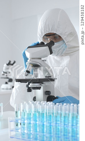 Medical scientist in protective suit using microscope while analyzing samples, conducting research in sterile laboratory environment. Close-up of test tubes with blue liquid 129438182