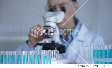 A female scientist, wearing a lab coat and safety glasses, is adjusting a binocular microscope surrounded by test tubes in laboratory setting. Medicine and science 129438183