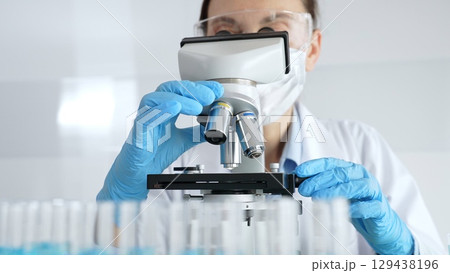 Woman scientist wearing protective gear is checking microscope, analyzing blue test tubes in modern laboratory research setting. Medicine, healthcare and science concept Woman scientist wearing protective gear is checking microscope, analyzing blue test tubes in modern laboratory research setting. Medicine, healthcare and science concept 129438196