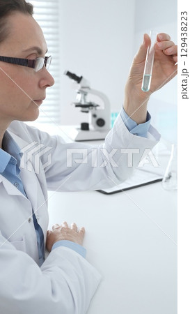 Female researcher examining clear liquid in test tube, sitting near microscopes and lab equipment within sterile scientific workspace. Medicine, healthcare and science concept Female researcher examining clear liquid in test tube, sitting near microscopes and lab equipment within sterile scientific workspace. Medicine, healthcare and science concept 129438223