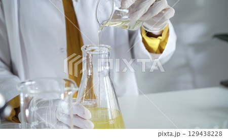 A scientist wearing a lab coat and white gloves is pouring a yellow oily liquid from one beaker to another in a laboratory setting, close up. Science and medicine 129438228