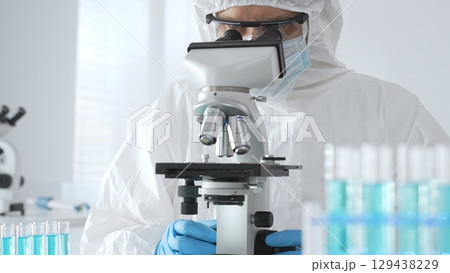 A male doctor in a protective suit and gloves uses a microscope while analyzing samples, conducting research in a laboratory. Close-up of test tubes with blue liquid A male doctor in a protective suit and gloves uses a microscope while analyzing samples, conducting research in a laboratory. Close-up of test tubes with blue liquid 129438229