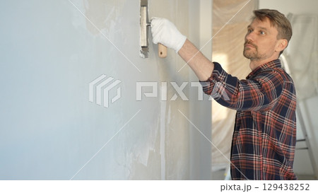 Male construction worker wearing gloves is carefully applying plaster to a wall using a hand tool, contributing to a home renovation or construction project Male construction worker wearing gloves is carefully applying plaster to a wall using a hand tool, contributing to a home renovation or construction project 129438252