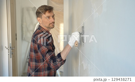 Male construction worker wearing gloves is carefully applying plaster to a wall using a hand tool, contributing to a home renovation or construction project Male construction worker wearing gloves is carefully applying plaster to a wall using a hand tool, contributing to a home renovation or construction project 129438271