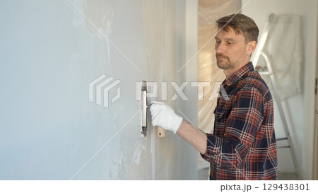 Male construction worker wearing gloves is carefully applying plaster to a wall using a hand tool, contributing to a home renovation or construction project Male construction worker wearing gloves is carefully applying plaster to a wall using a hand tool, contributing to a home renovation or construction project 129438301