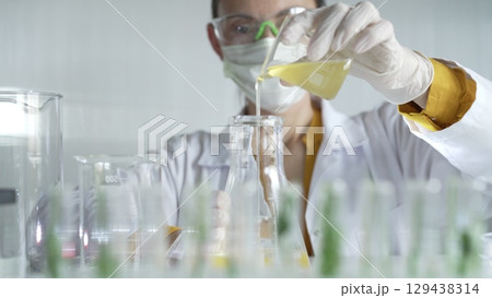 Woman scientist wearing a lab coat, white gloves, and a mask is pouring a yellow oily liquid from a beaker into an Erlenmeyer flask in a laboratory setting, close up. Science and medicine 129438314