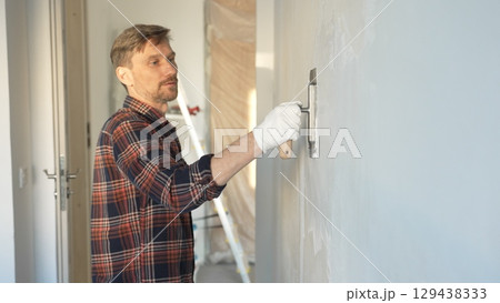 Male construction worker wearing protective gloves smoothing plaster onto wall surface during home renovation using professional trowel tool 129438333