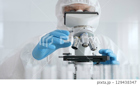 Male scientific researcher or virologist wearing protective gear adjusting microscope, analyzing lab samples with precision in sterile research environment. Medicine concept 129438347