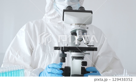 A male doctor in a protective suit and gloves uses a microscope while analyzing samples, conducting research in a laboratory. Close-up of test tubes with blue liquid 129438352