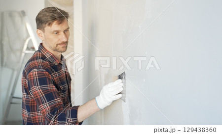 Construction worker man wearing gloves and holding a putty knife is applying plaster on a white wall, performing home renovation and improvement Construction worker man wearing gloves and holding a putty knife is applying plaster on a white wall, performing home renovation and improvement 129438360