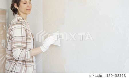 Woman applying plaster to wall with putty knife, wearing protective gloves and creating smooth surface during home renovation work Woman applying plaster to wall with putty knife, wearing protective gloves and creating smooth surface during home renovation work 129438368
