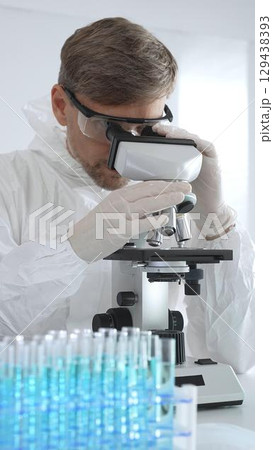 Male virologist in medical gown and gloves examines virus samples under microscope while conducting research in sterile laboratory conditions. Close-up of test tubes with blue liquid 129438393