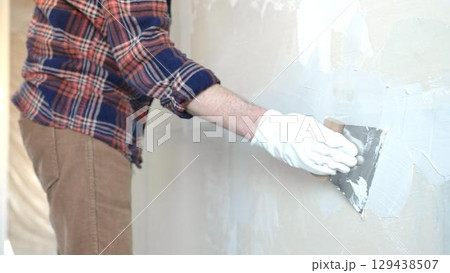 Unknown male construction worker wearing protective gloves smoothing plaster on interior wall during home renovation, applying material with professional trowel technique, close-up Unknown male construction worker wearing protective gloves smoothing plaster on interior wall during home renovation, applying material with professional trowel technique, close-up 129438507