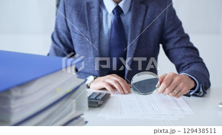 Professional businessman examining financial documents, magnifying glass in hand, calculator positioned on sleek desk workspace. Audit and taxes in business 129438511