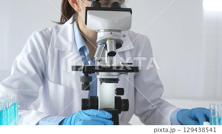 Female scientist wearing protective gear carefully adjusting microscope lens, analyzing medical research samples with precision in sterile laboratory environment Female scientist wearing protective gear carefully adjusting microscope lens, analyzing medical research samples with precision in sterile laboratory environment 129438541