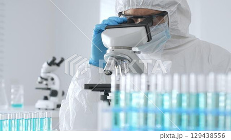 Male doctor in protective suit and gloves using microscope while analyzing samples while conducting research in sterile laboratory. Close-up of test tubes with blue liquid 129438556