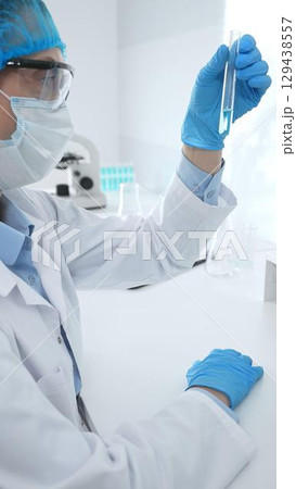 Medical science researcher wearing blue protective gear of cap, gloves and googles, is examining liquid in test tube in laboratory workspace. Medicine and health care concept 129438557